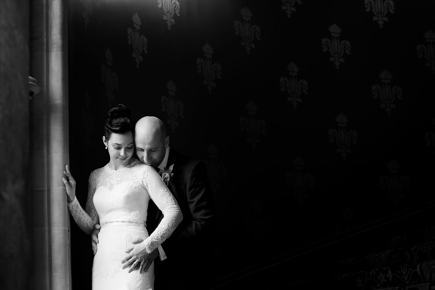 Intimate black and white wedding portrait of bride in lace gown and groom embracing near a patterned wall.