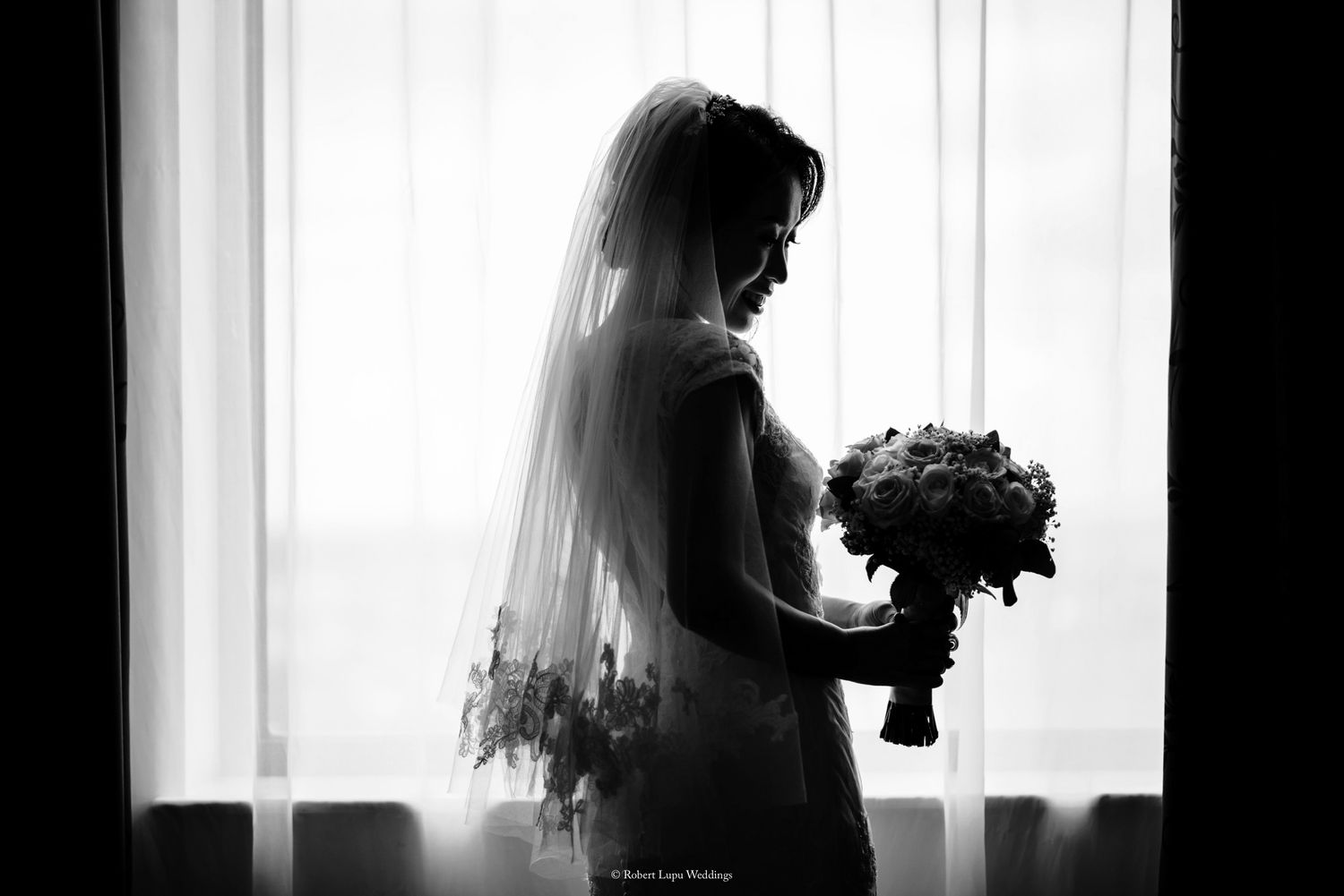 Black and white silhouette of a bride in veil holding bouquet standing by bright window on wedding day.