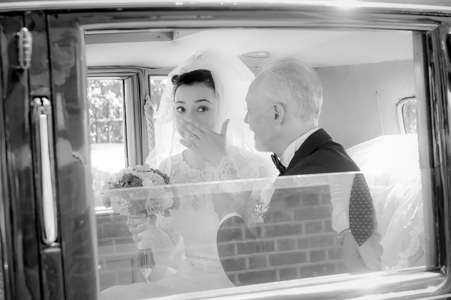 Black and white photo of a bride covering her mouth in surprise beside an older man, viewed through a car window.