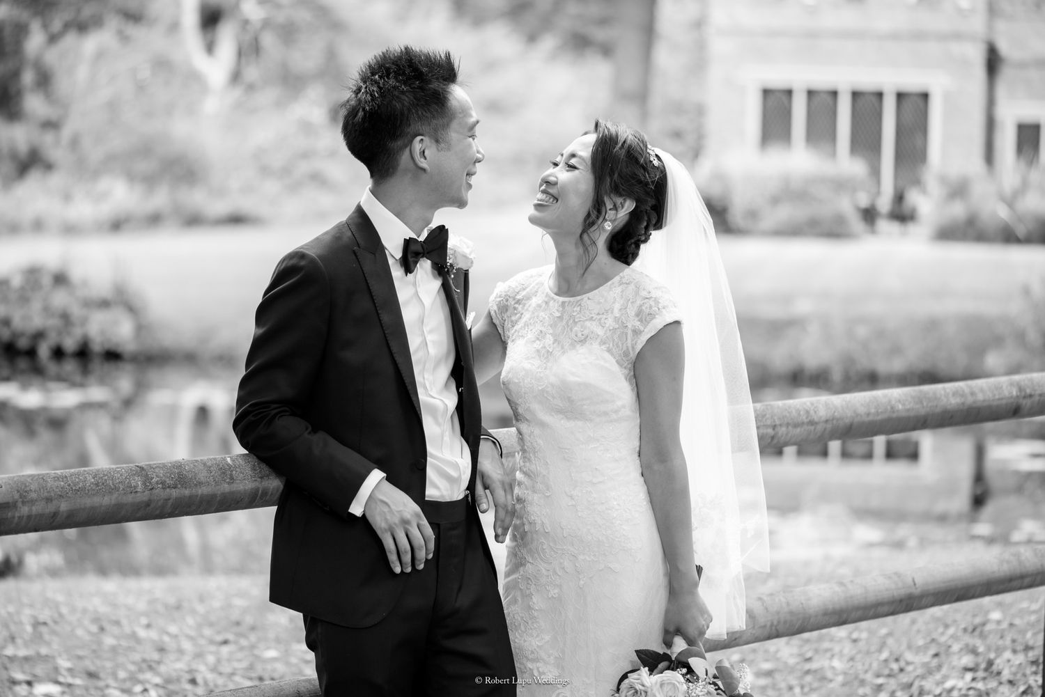 Black and white wedding photo of a smiling couple in formal attire standing by a wooden fence near a pond.