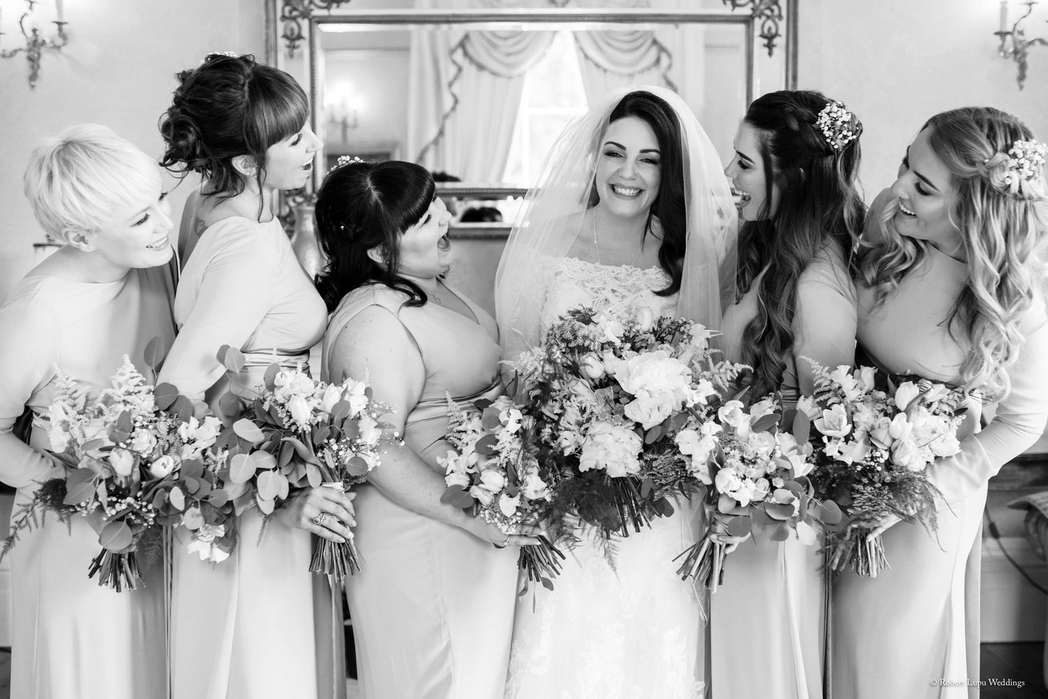 Black and white photo of a bride in a veil laughing with five bridesmaids in matching dresses holding floral bouquets.