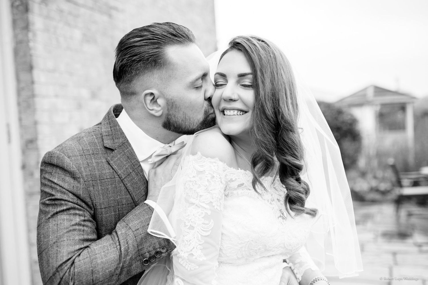 Black and white wedding photo of groom kissing bride on cheek, bride smiling in lace dress and veil.