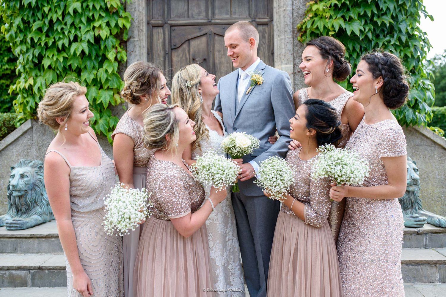 Groom in gray suit surrounded by bridesmaids in blush sequin dresses holding baby's breath bouquets at elegant wedding.