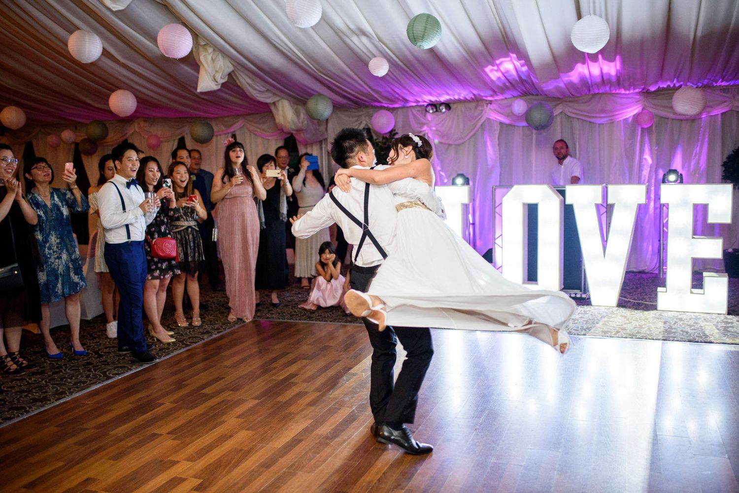 Groom lifts bride on dance floor at wedding reception with LOVE sign and balloon decorations.