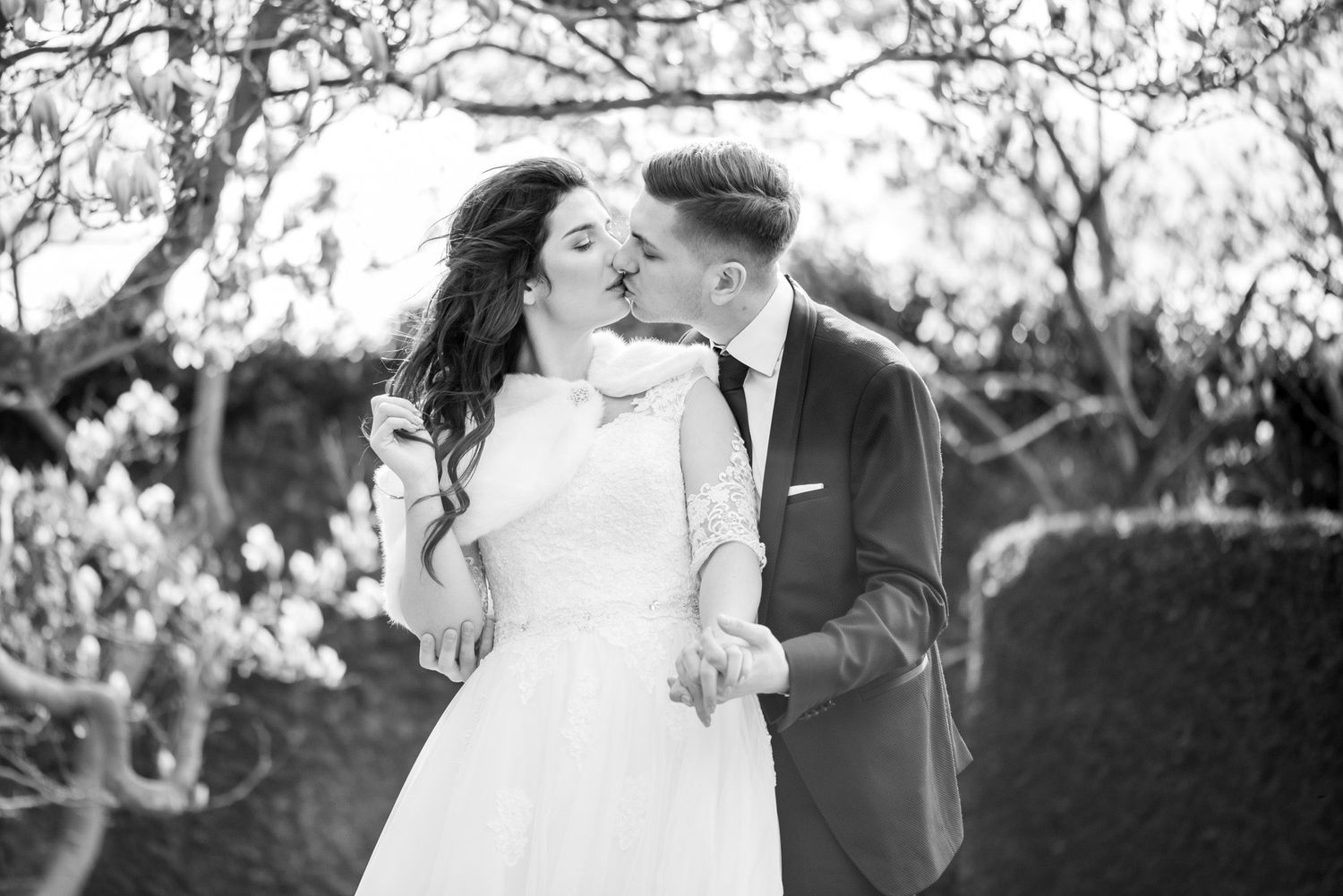 Black and white photo of a bride and groom sharing a kiss outdoors among blooming trees on their wedding day.