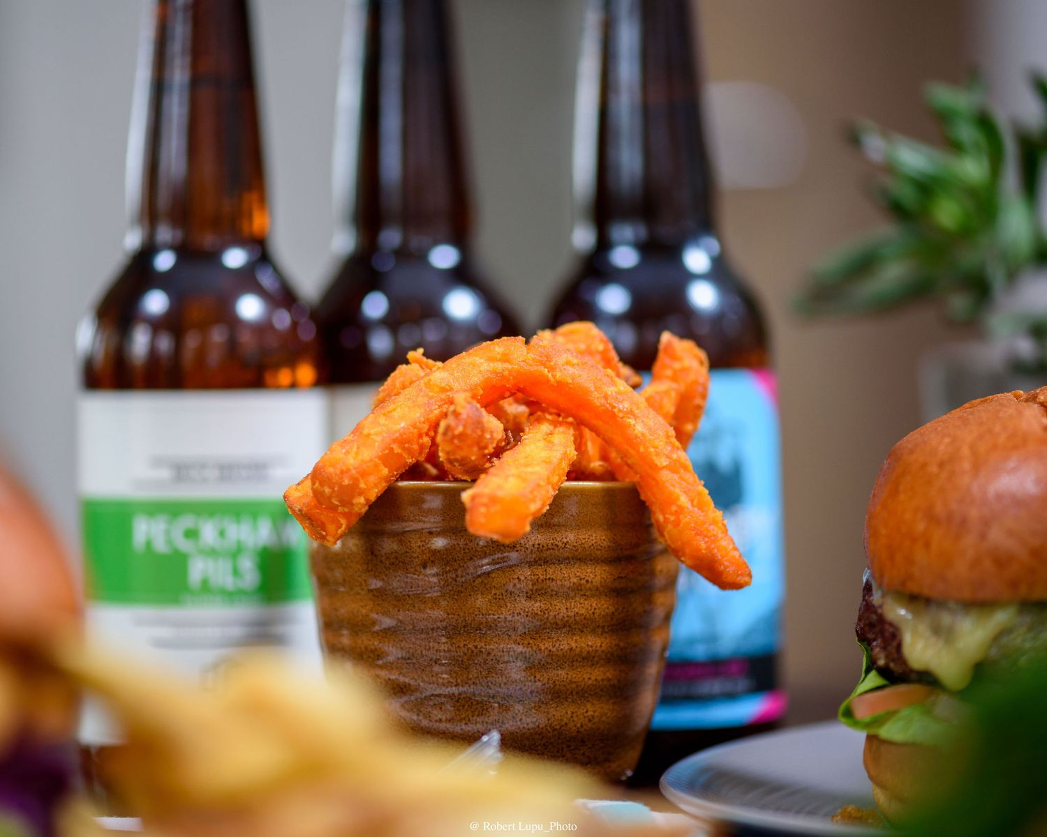 Sweet potato fries in a bowl with Peckham Pils beer bottles and a burger in the background.