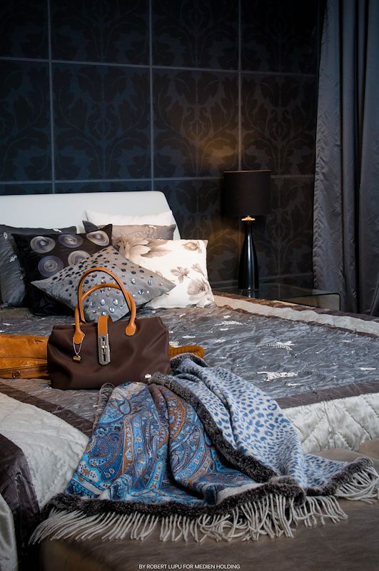 Elegant bedroom with blue damask walls, luxury bedding, a brown leather handbag with orange handles, and a blue paisley throw blanket.