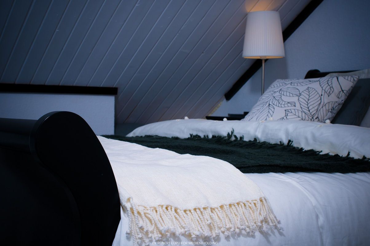 Cozy attic bedroom with white bedding, green throw blanket, decorative pillows, and a soft glowing lamp at night.