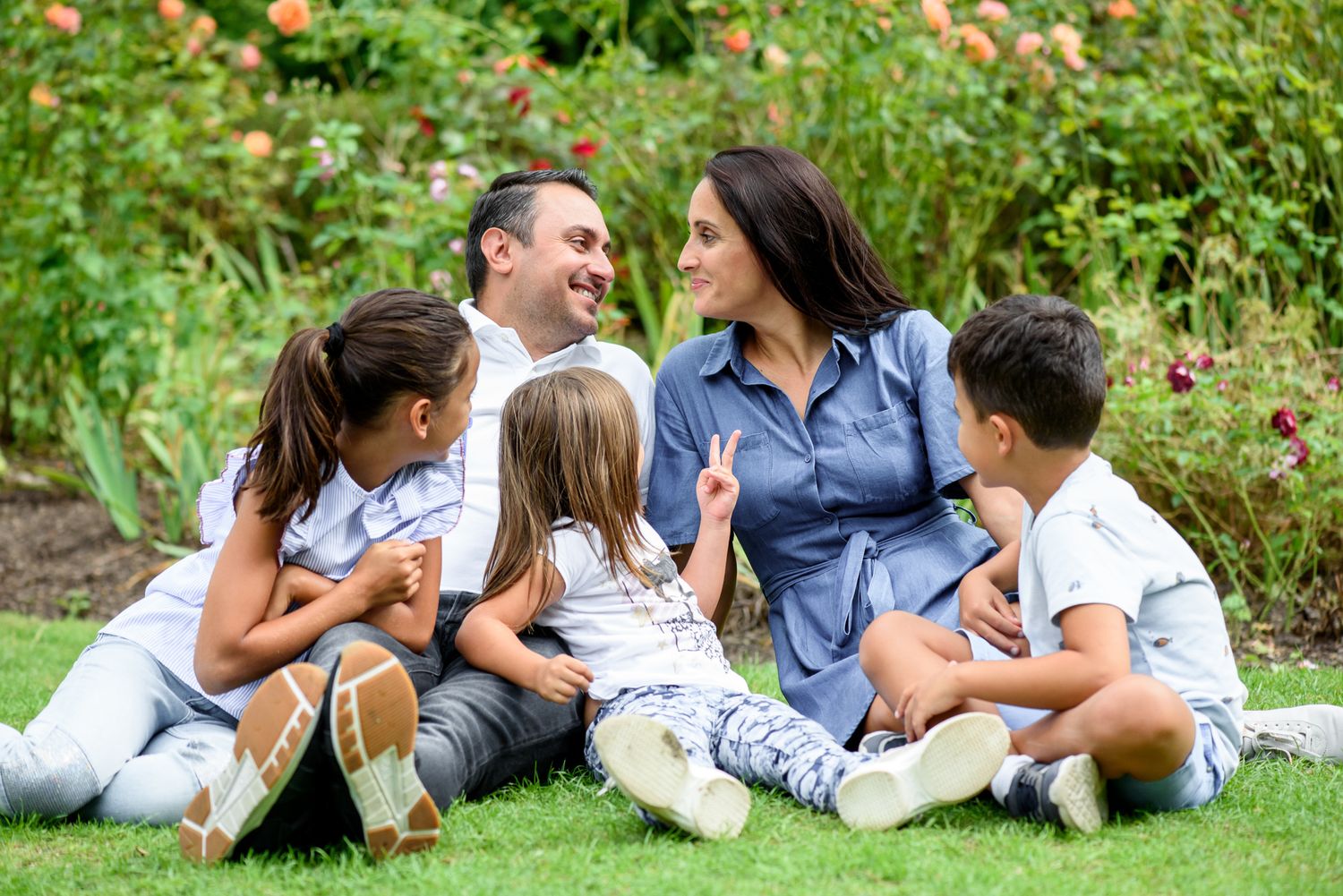 Happy family of five sitting on grass in a garden, parents and three children laughing together outdoors.