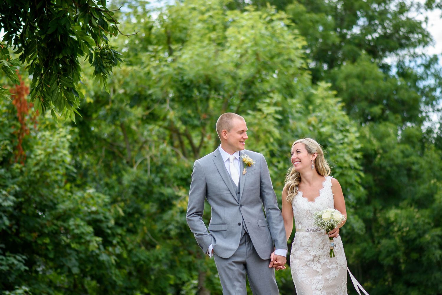 Bride and groom holding hands walking outdoors in lush green garden, groom in gray suit, bride in white lace wedding dress.