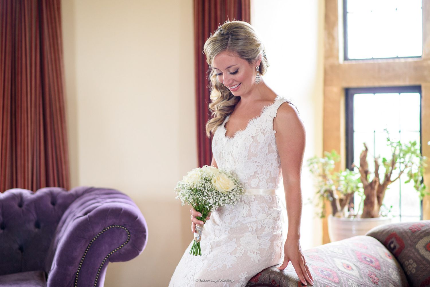 Bride in white lace gown holding bouquet of white flowers, seated on floral sofa near purple velvet chair.