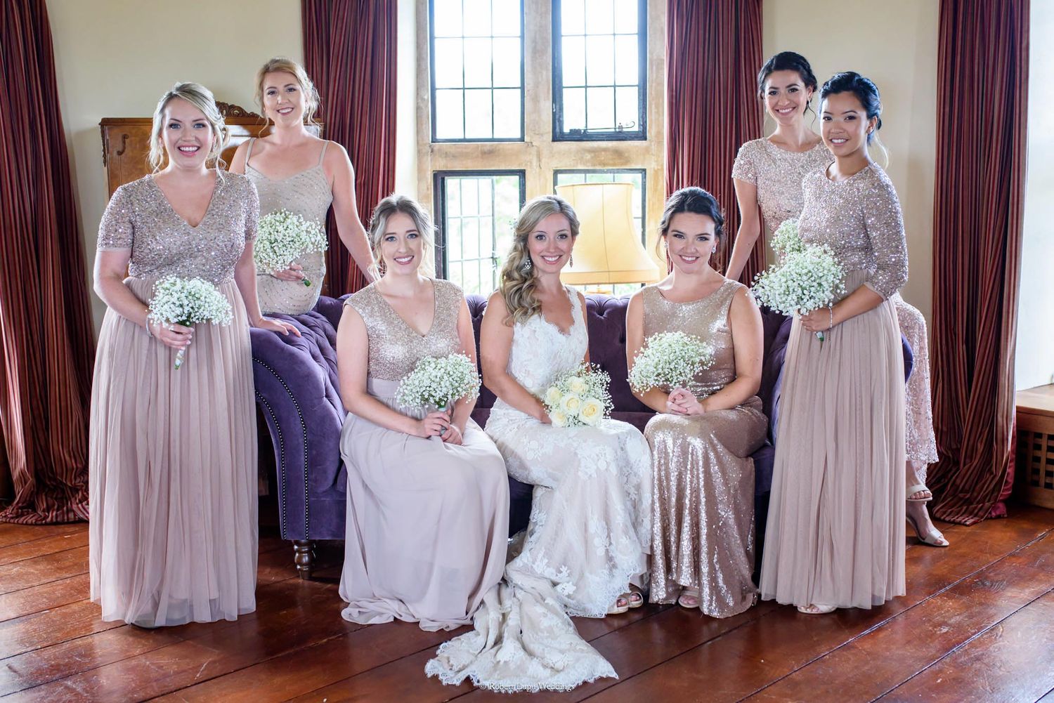 Bride and bridesmaids in blush and silver gowns holding white bouquets posing together in elegant venue.