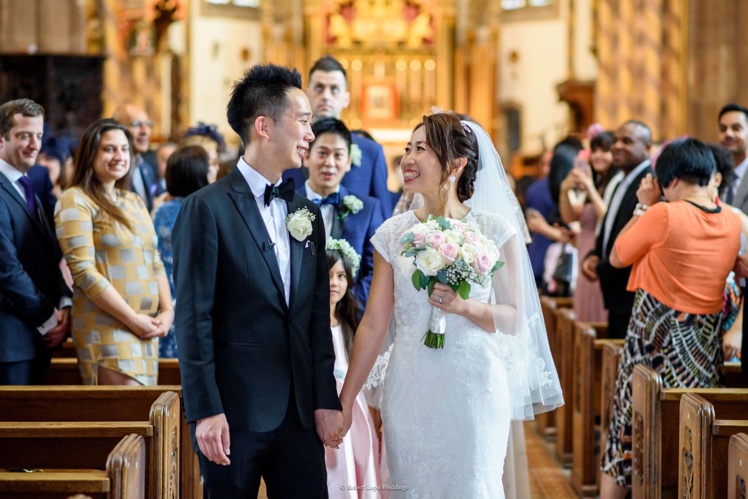 Smiling bride and groom walk down church aisle holding hands, bride carries pink and white bouquet, guests watch ceremony.
