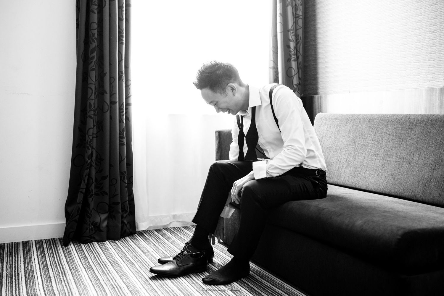 Black and white photo of a groom in suspenders sitting on a sofa, putting on dress shoes before his wedding.
