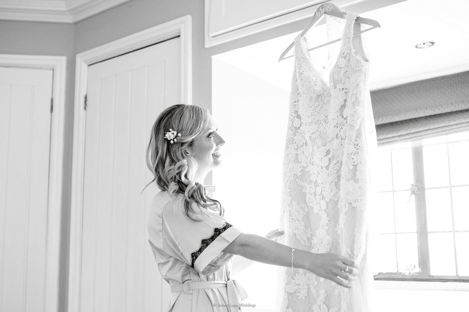 Bride in robe admiring lace wedding dress hanging by window in black and white photo.