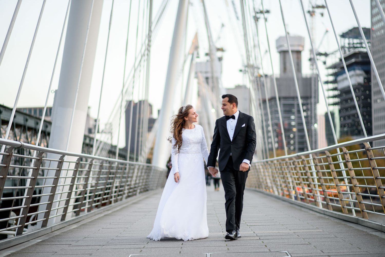 Bride and groom walking hand in hand on a bridge with city skyline backdrop on their wedding day.