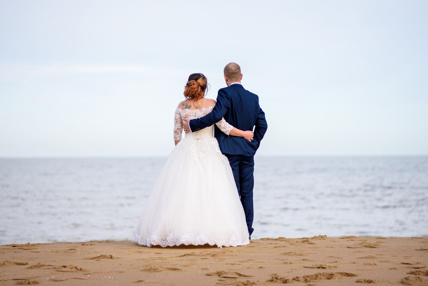 Bride and groom standing together on a sandy beach, gazing at the ocean, wearing wedding dress and navy suit.