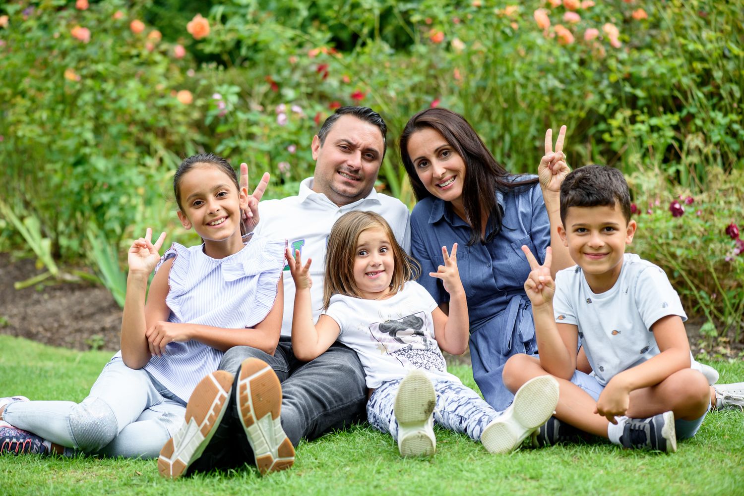 Smiling family of five posing with peace signs while sitting on grass in a colorful garden setting.