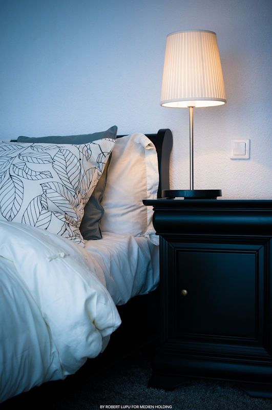 Cozy bedroom with white bedding, leaf-patterned pillows, dark wood nightstand, and glowing table lamp in soft blue light.