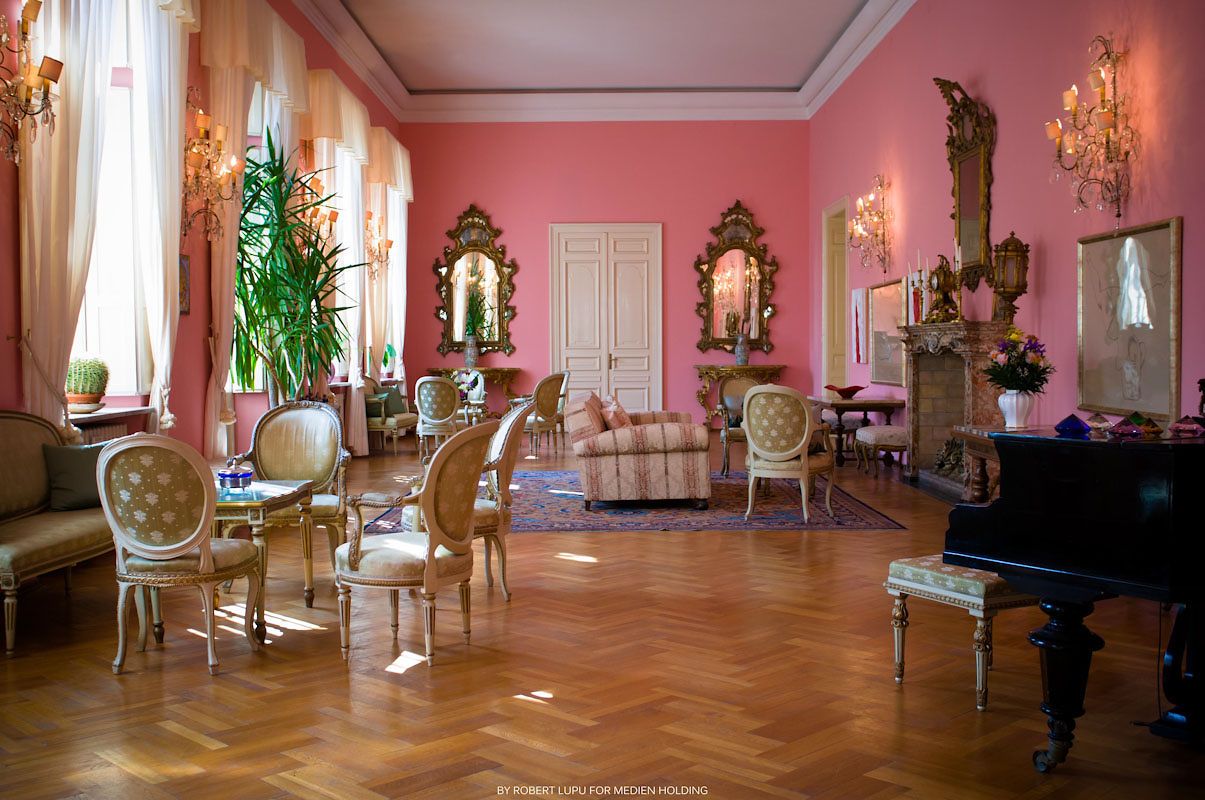 Elegant pink baroque salon with ornate gold mirrors, Louis XVI chairs, parquet floors, and a grand piano in a luxurious palace interior.