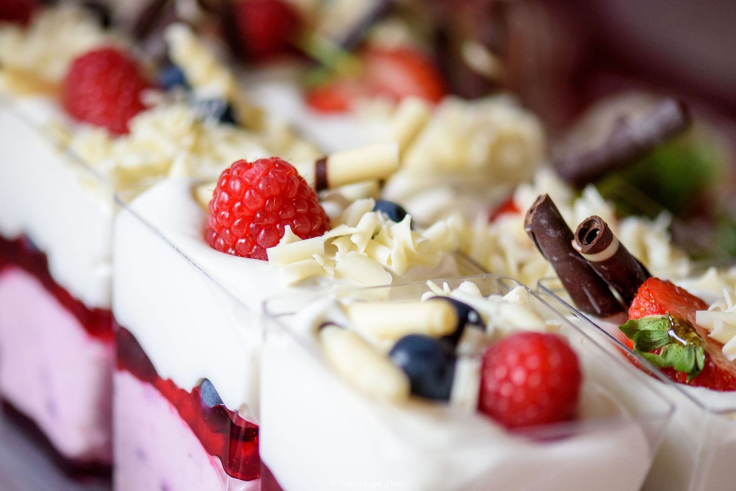 Elegant dessert slices topped with fresh raspberries, blueberries, white chocolate shavings, and chocolate sticks on creamy layers.