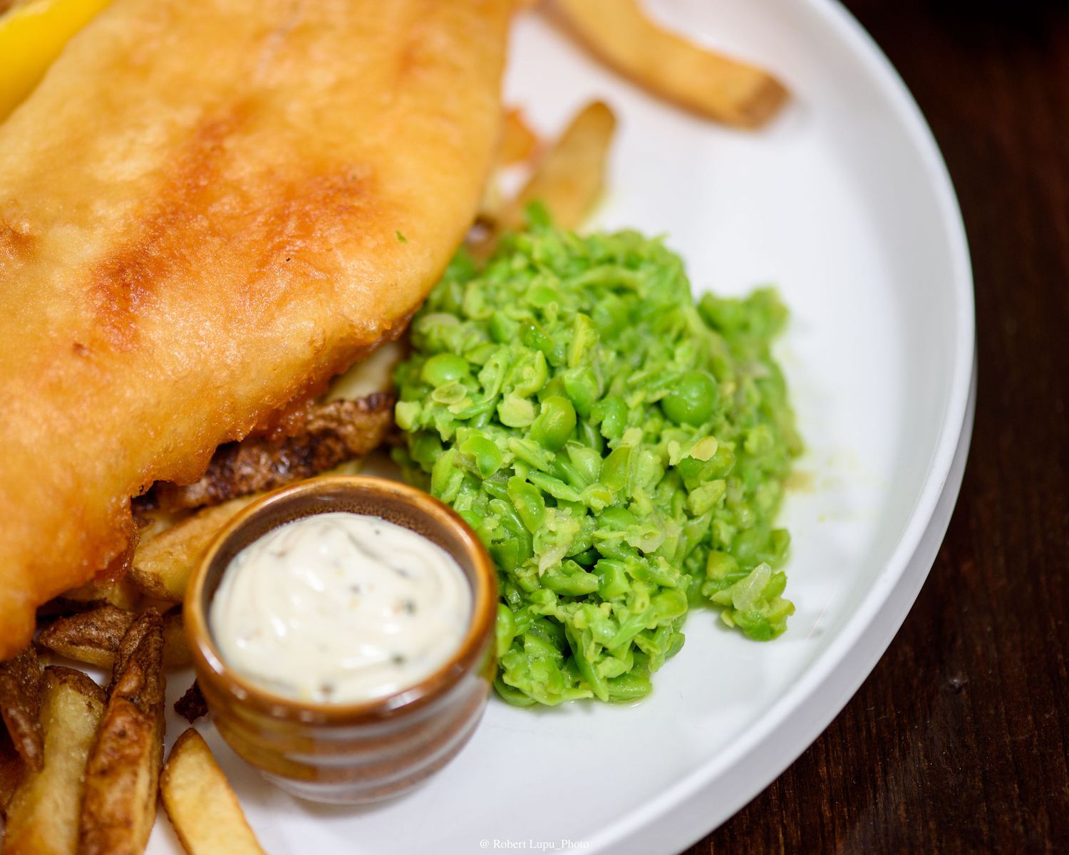 Classic fish and chips with golden battered fish, crispy fries, mushy peas, and tartar sauce on a white plate.