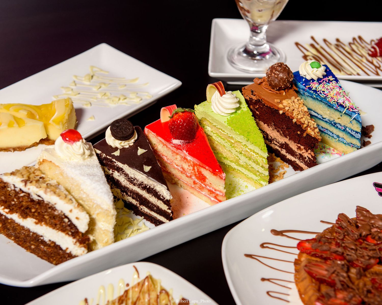 Assorted colorful cake slices on a white platter, featuring strawberry, matcha, chocolate, and rainbow layers at a dessert restaurant.