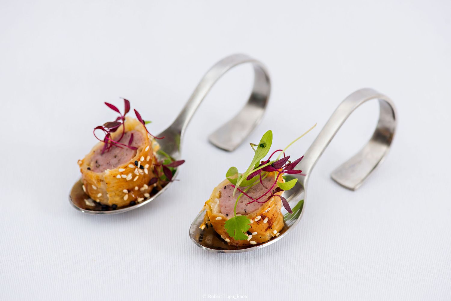 Two elegant silver tasting spoons with tuna tartare, sesame seeds, and micro greens on white background.