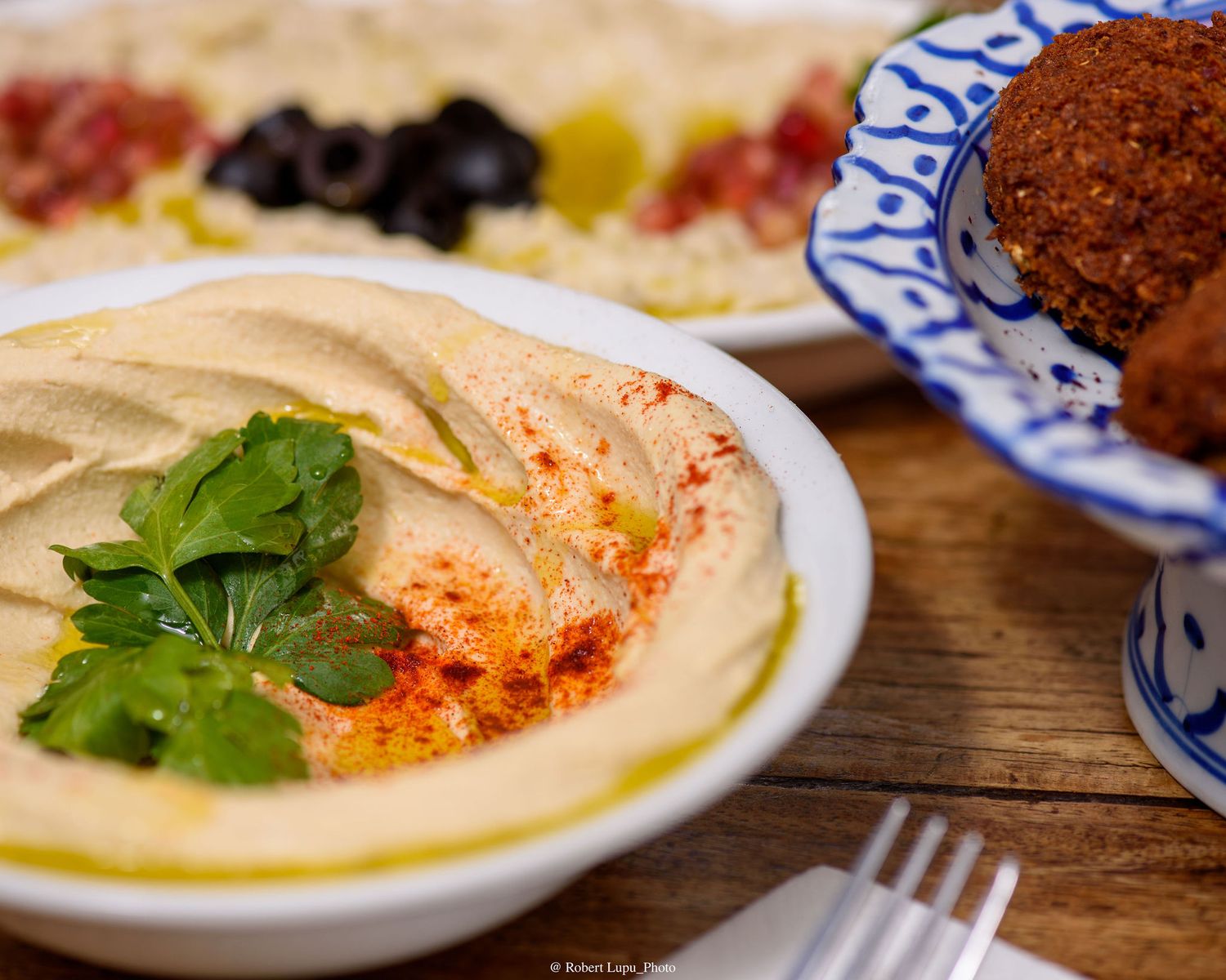 Creamy hummus garnished with paprika and parsley, served with falafel and olives on a rustic wooden table.