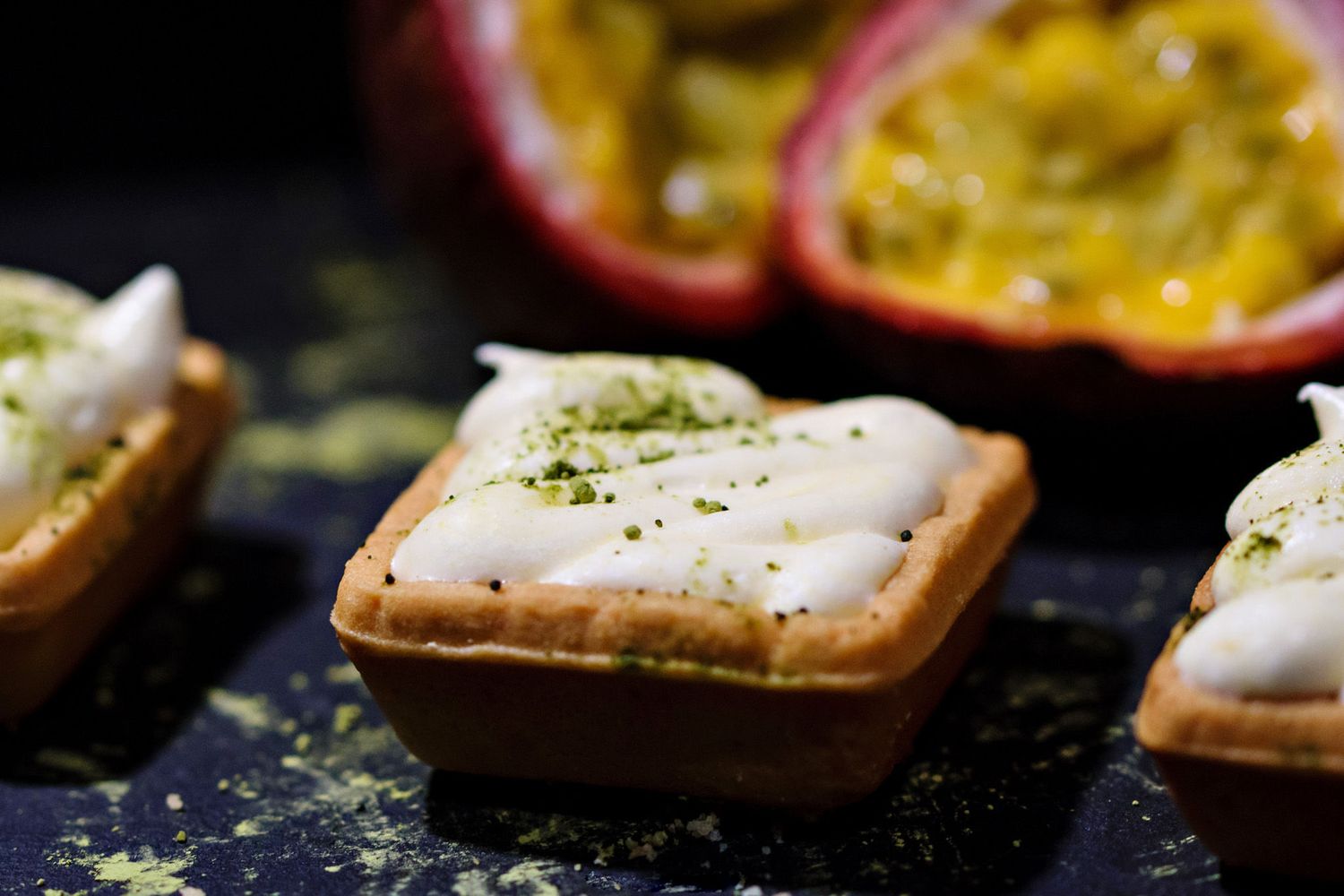 Square pastry tarts filled with cream and garnished with green zest, beside a halved passion fruit on a dark slate.