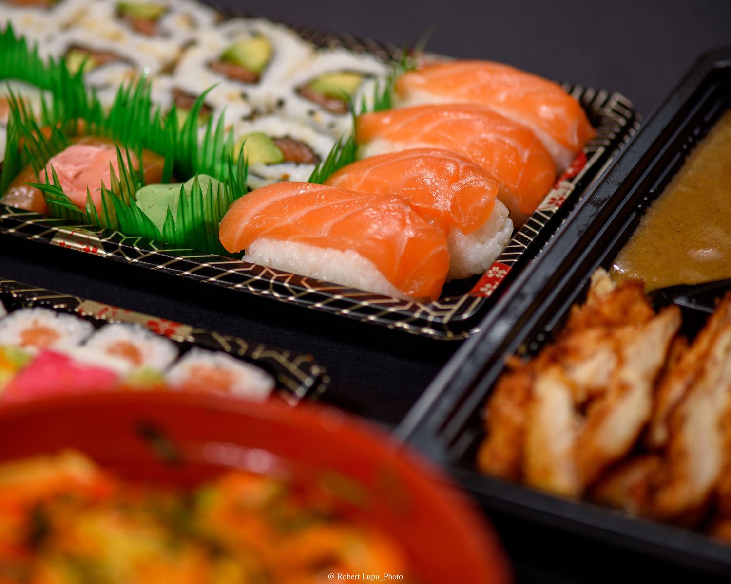 Close-up of a Japanese bento box with salmon nigiri sushi, maki rolls, and grilled chicken on a dark tray.