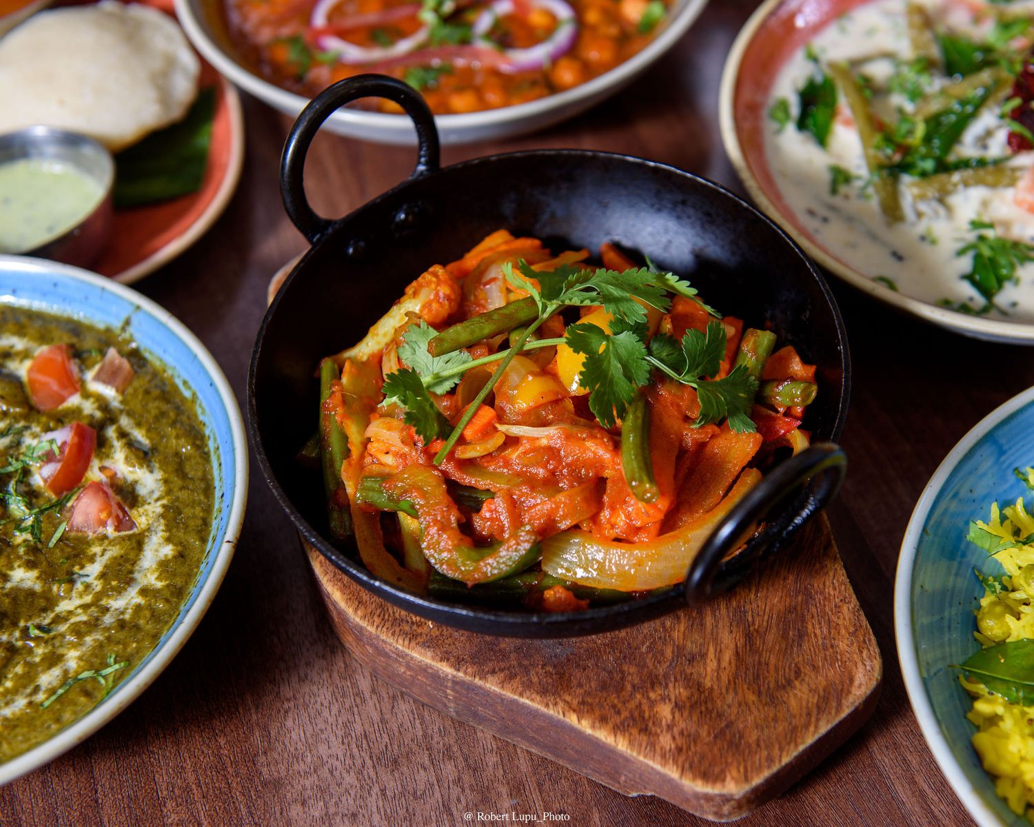 Colorful Indian vegetable curry served in a black karahi on a wooden board surrounded by side dishes.