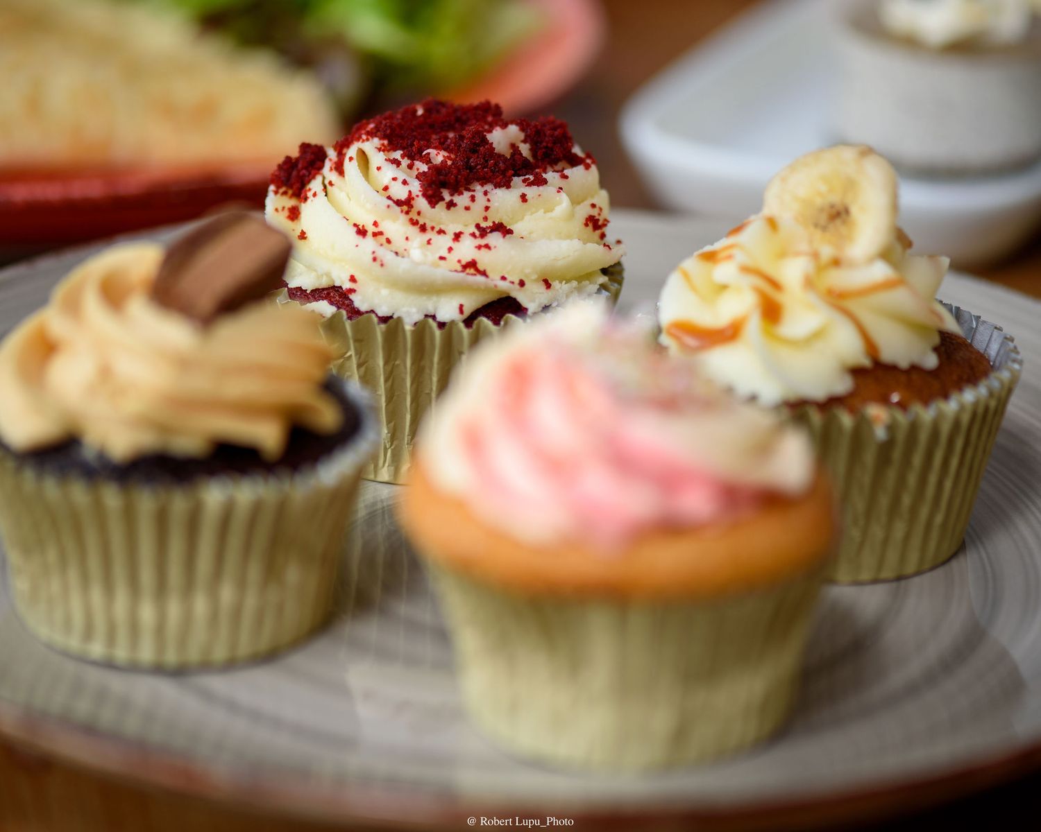 Four assorted frosted cupcakes on a plate, featuring red velvet, banana, strawberry, and vanilla flavors.