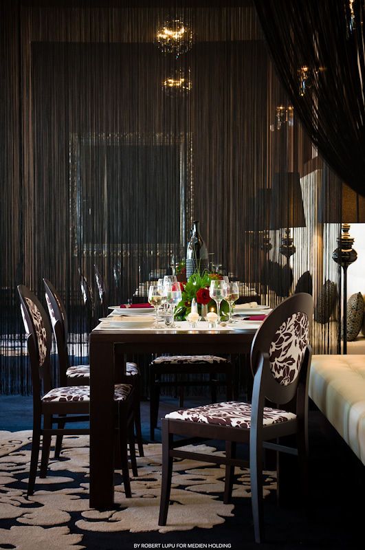 Elegant dark dining room with black curtains, floral chairs, set table with wine glasses and bottles, chandelier overhead.