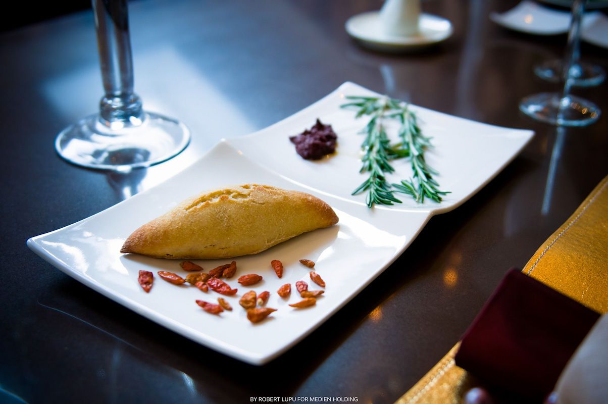 Gourmet bread roll served on white rectangular plate with rosemary, goji berries, and red sauce.