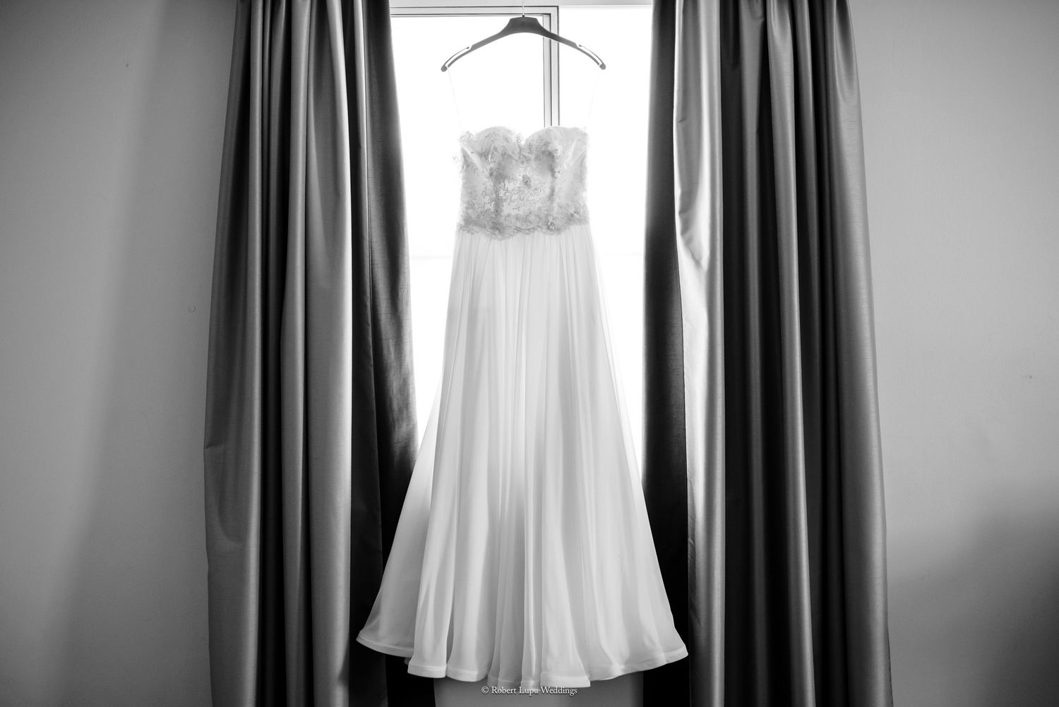 Black and white photo of an elegant strapless wedding gown with lace bodice hanging on a curtained window.