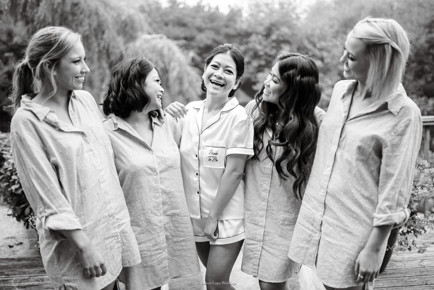 Five bridesmaids in matching linen robes laughing together outdoors in a black and white wedding photo.