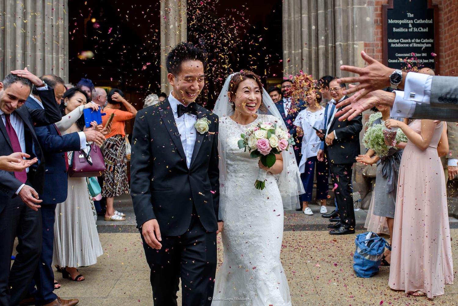 Asian couple exits church in confetti shower, bride in lace gown holding bouquet, groom in tuxedo, guests celebrating.