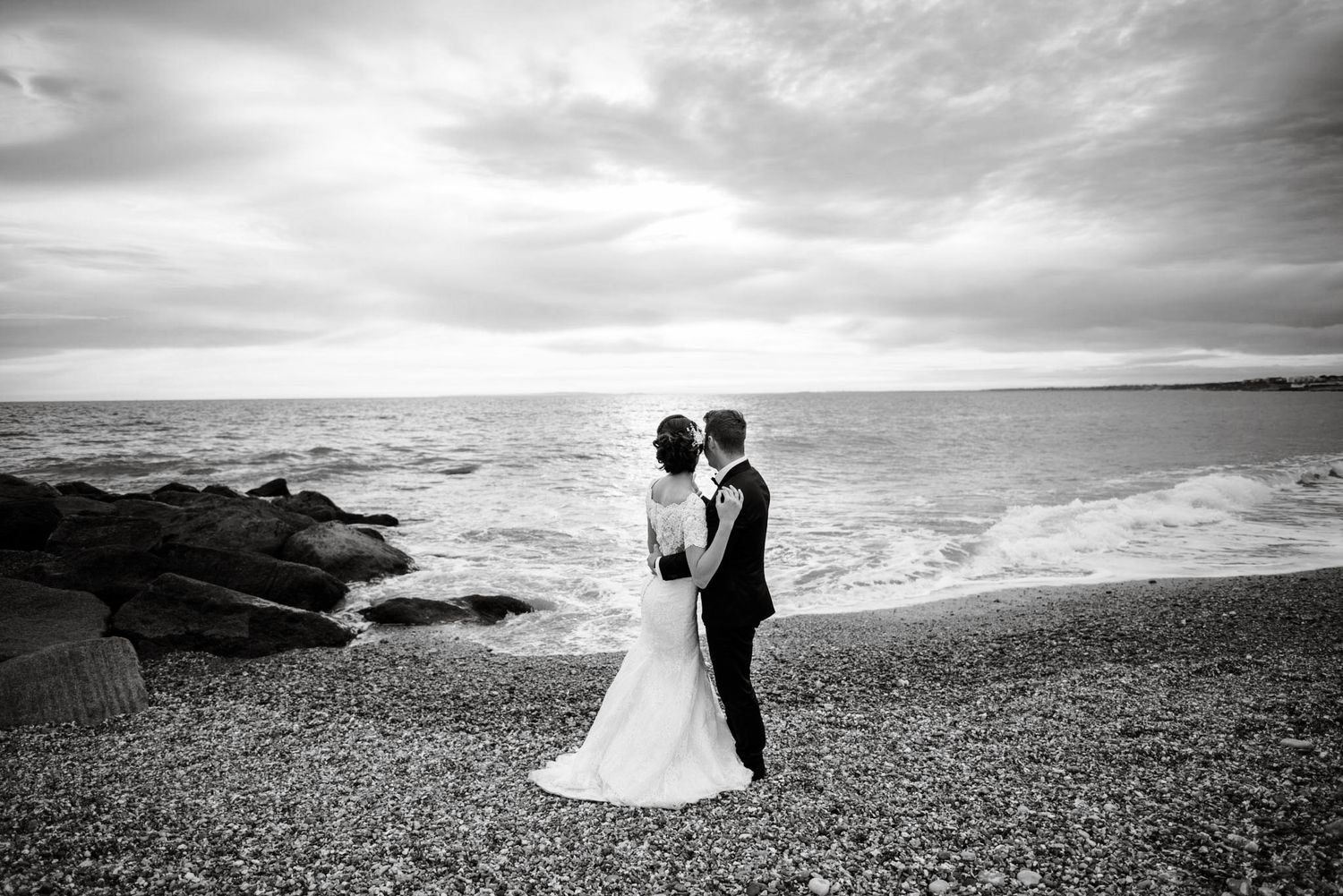 Bride and groom share a romantic moment on a rocky beach by the ocean in a black and white wedding photo.