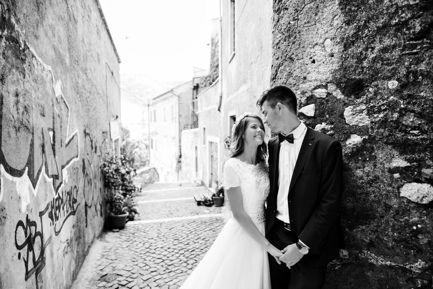 Bride and groom share a romantic moment in a cobblestone alley with graffiti walls in a black and white wedding photo.