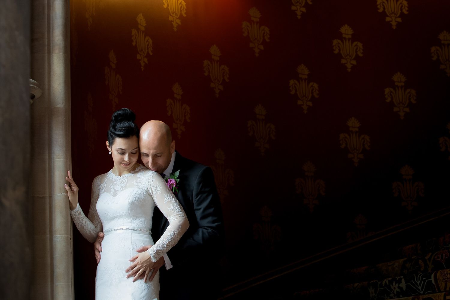 Bride and groom share an intimate moment leaning together against ornate red wall with gold fleur-de-lis pattern.