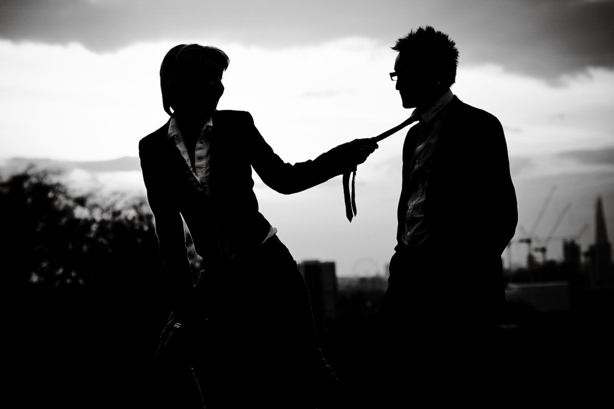 Black and white silhouette of two people in suits, one grabbing the other's tie outdoors.