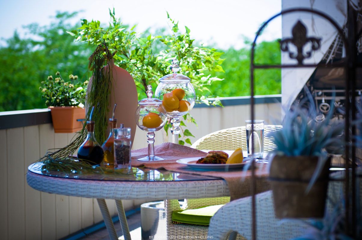 Elegant balcony table with wine glasses, fresh fruits, and lush green plants on a sunny outdoor terrace.