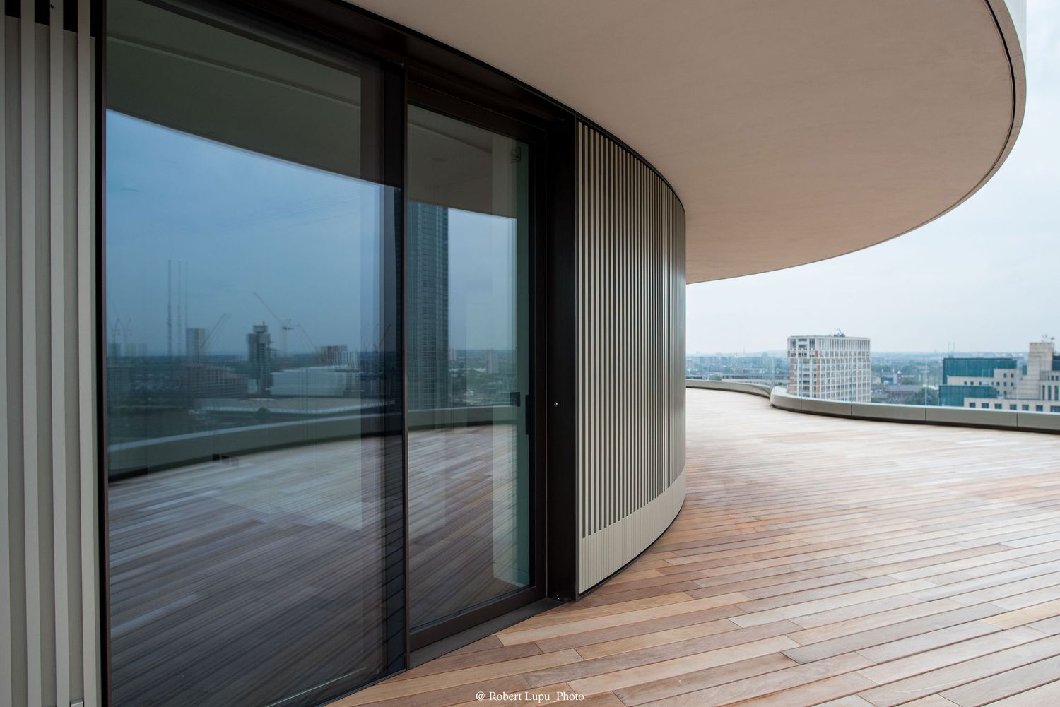 Modern curved building rooftop terrace with wooden deck flooring and floor-to-ceiling glass sliding doors.