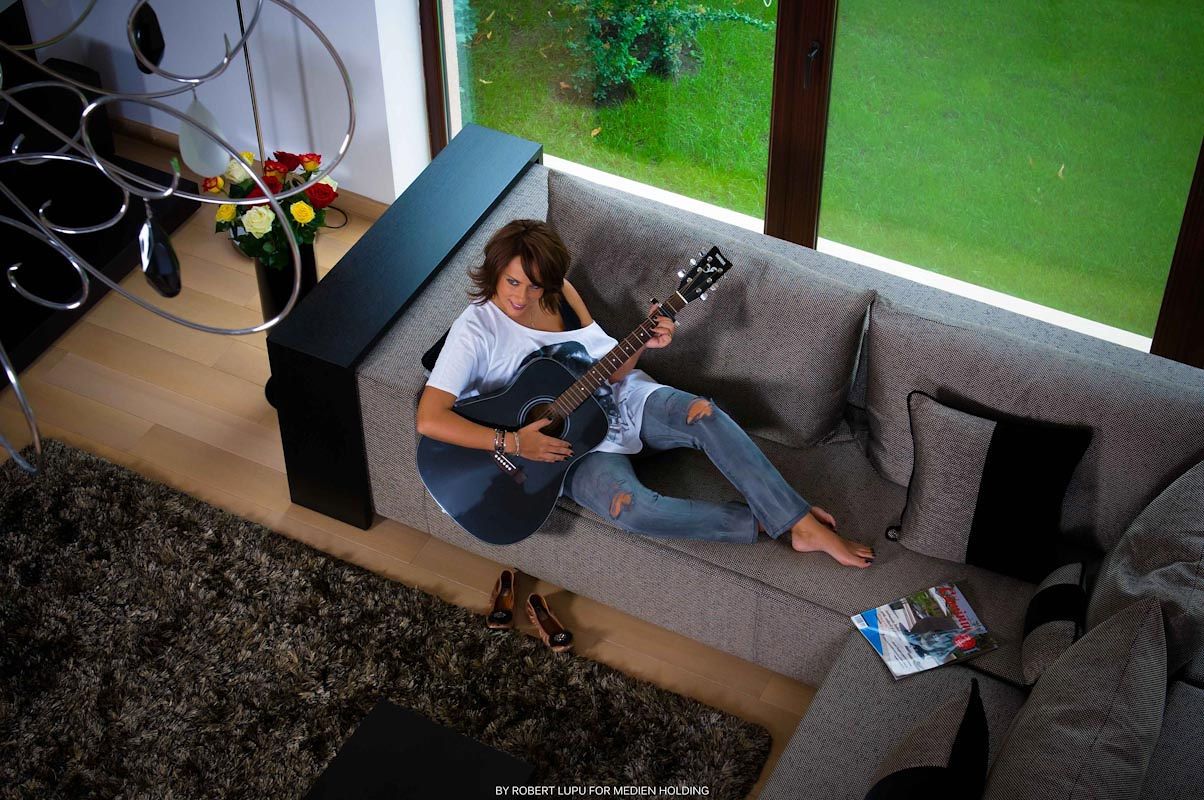 Aerial view of a woman playing acoustic guitar on a gray sofa in a modern living room.