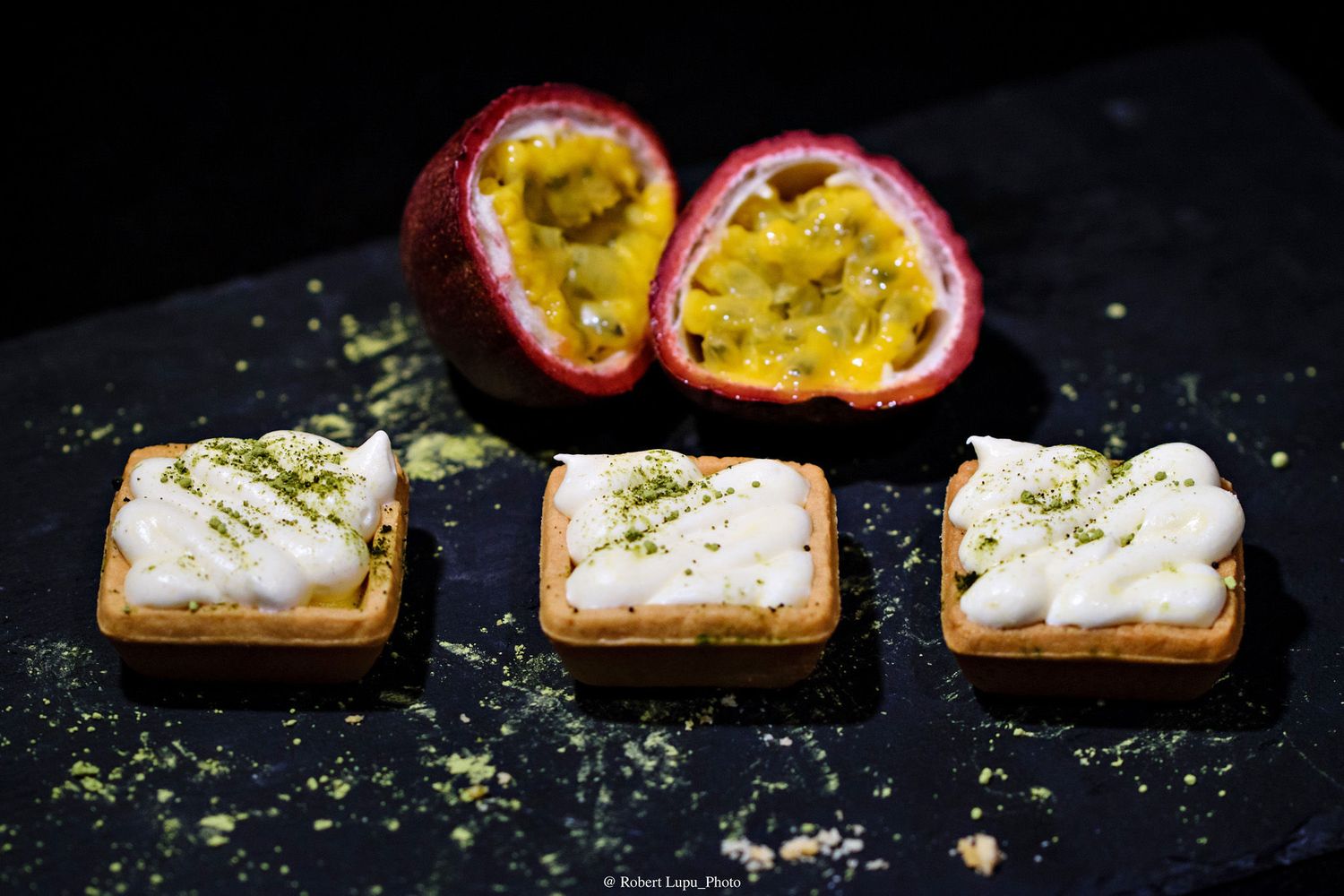 Three square passion fruit tarts with white meringue topping beside halved fresh passion fruits on dark slate.