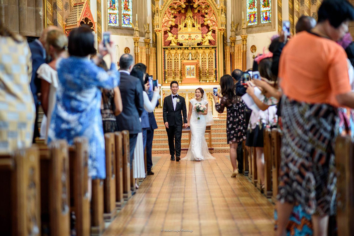 Bride and groom walk down the aisle of an ornate church surrounded by wedding guests.