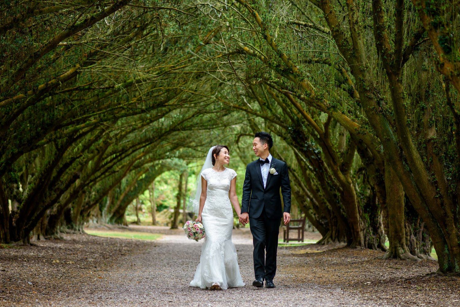 Bride and groom walk hand-in-hand along a tree-lined path, smiling at each other on their wedding day.