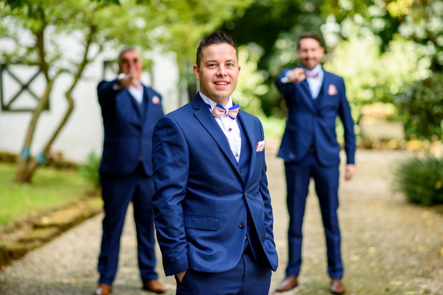 Three groomsmen in navy blue suits pose outdoors at a wedding, with the groom standing prominently in the foreground.