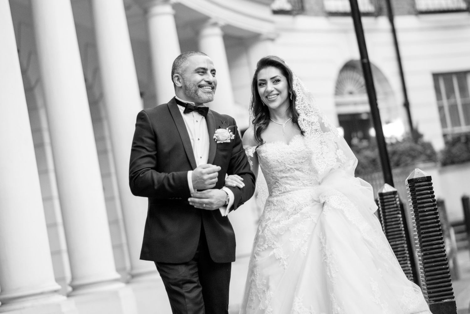 Black and white photo of a bride and groom walking together near elegant columns on their wedding day.